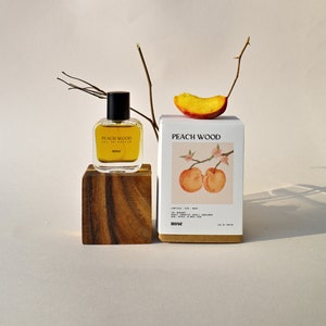 May include: A small bottle of Peach Wood Eau de Parfum by nose. The bottle is sitting on a wooden block. The bottle is clear and contains a yellow liquid. The bottle has a black cap. The bottle is in front of a white box with the words "Peach Wood" printed on it. The box has a picture of two peaches on it. The peaches are red and yellow and have a green stem.