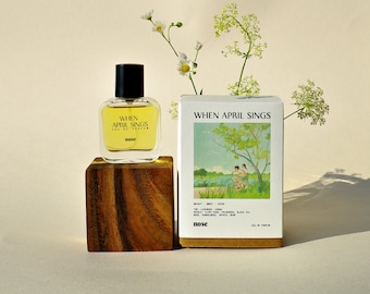 May include: A clear glass bottle of perfume with a yellow liquid. The bottle is labeled "WHEN APRIL SINGS" and "EAU DE PARFUM". The bottle is sitting on a wooden block. The background is a white wall with a bouquet of white flowers.