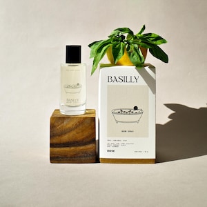 May include: A white box with the text "BASILLY" and a drawing of a bathtub with a person inside. The box is labeled "ROOM SPRAY" and has a list of ingredients. The box is sitting on a wooden surface next to a clear glass bottle with a black cap. The bottle is labeled "BASILLY ROOM SPRAY".