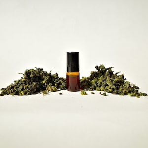 May include: A small brown glass bottle with a black roller ball top, surrounded by green tea leaves on a white background.