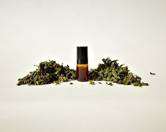 May include: A small brown glass bottle with a black roller ball top, surrounded by green tea leaves on a white background.