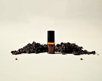 May include: A small brown glass bottle with a black cap, filled with a dark liquid, sits on a white surface surrounded by loose black tea leaves.