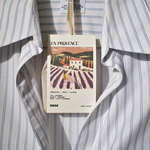 May include: A white and blue striped shirt with a small white tag attached. The tag features a picture of a lavender field and a house with the text "EN PROVENCE" and "SCENT SACHET". The tag also lists the scent notes: "HERBACEOUS | FRESH | CALMING", "TOP: LAVENDER", "MIDDLE: ROSEMARY", "BASE: ATLAS CEDARWOOD", and "nose".