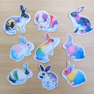 Puede incluir: Una colección de nueve pegatinas de conejos coloridos. Cada pegatina presenta un diseño único de acuarela de un conejo en varias poses y combinaciones de colores, incluyendo patrones de arcoíris, pastel y moteados. Las pegatinas están sobre una superficie de madera clara.