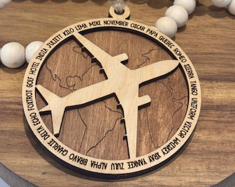 Könnte beinhalten: Ein Holzschmuckstück in Form eines Flugzeugs, das über einer Weltkarte fliegt. Der Schmuck ist mit dem NATO-Phonetischen Alphabet graviert, beginnend mit "Alpha" und endend mit "Tango".