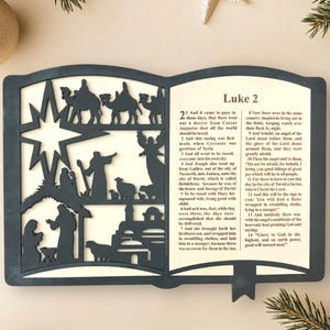 May include: A wooden book-shaped ornament with a black silhouette of the nativity scene and a portion of the Bible verse Luke 2:1-14. The ornament is a Christmas decoration.