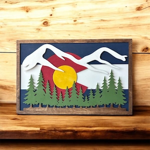 May include: A rectangular wooden wall art piece featuring a layered design of the Colorado state flag. The scene includes white mountain silhouettes, a central yellow sun, and a row of green pine trees, all enclosed within a dark wood frame.