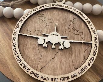 Könnte beinhalten: Ein Holzschmuckstück in Form einer Weltkarte mit einem Flugzeug in der Mitte. Der Schmuck ist mit dem NATO-Phonetischen Alphabet graviert, beginnend mit "Alpha" und endend mit "Zulu".