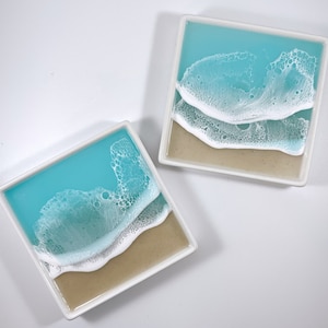 May include: Two square ceramic coasters with a beach scene. The coasters feature a light blue ocean with white foamy waves crashing on a beige sandy beach.