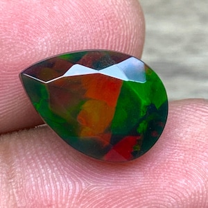 Puede incluir: Una piedra preciosa de ópalo negro en forma de lágrima con un brillo iridiscente verde, rojo y naranja.