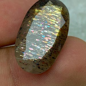 Op de afbeelding: Een grote, ovale edelsteen met een bruine, iriserende en glinsterende uitstraling. De edelsteen heeft een facetgeslepen vorm en toont een regenboog aan kleuren.