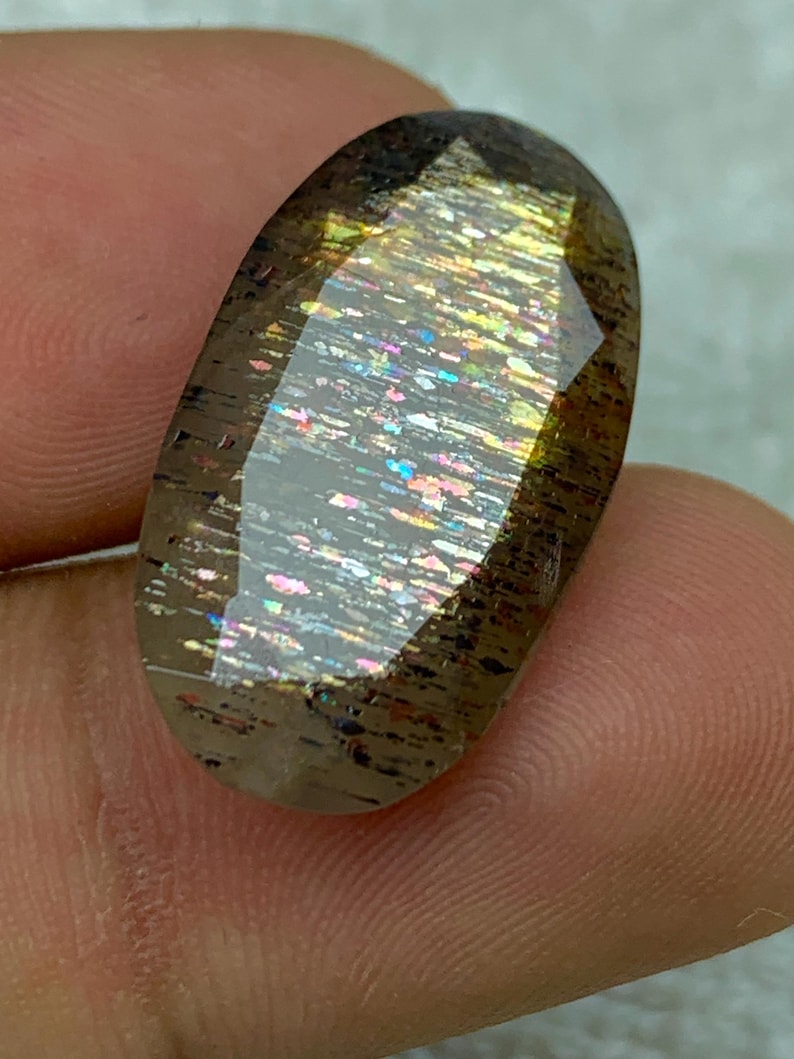 Op de afbeelding: Een grote, ovale edelsteen met een bruine, iriserende en glinsterende uitstraling. De edelsteen heeft een facetgeslepen vorm en toont een regenboog aan kleuren.