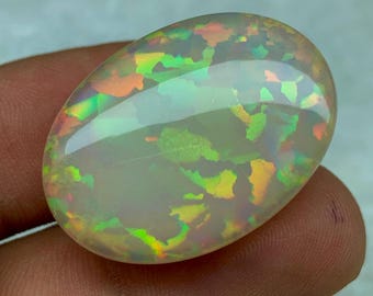 May include: A white opal cabochon with a green, orange, and red iridescent sheen. The opal is oval shaped and has a smooth, polished surface.