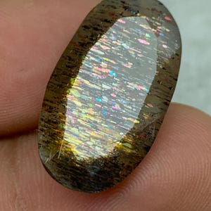 Op de afbeelding: Een grote, ovale edelsteen met een donkerbruine basis en een iriserende regenboogkleurige schittering. De edelsteen heeft een glad, gepolijst oppervlak.