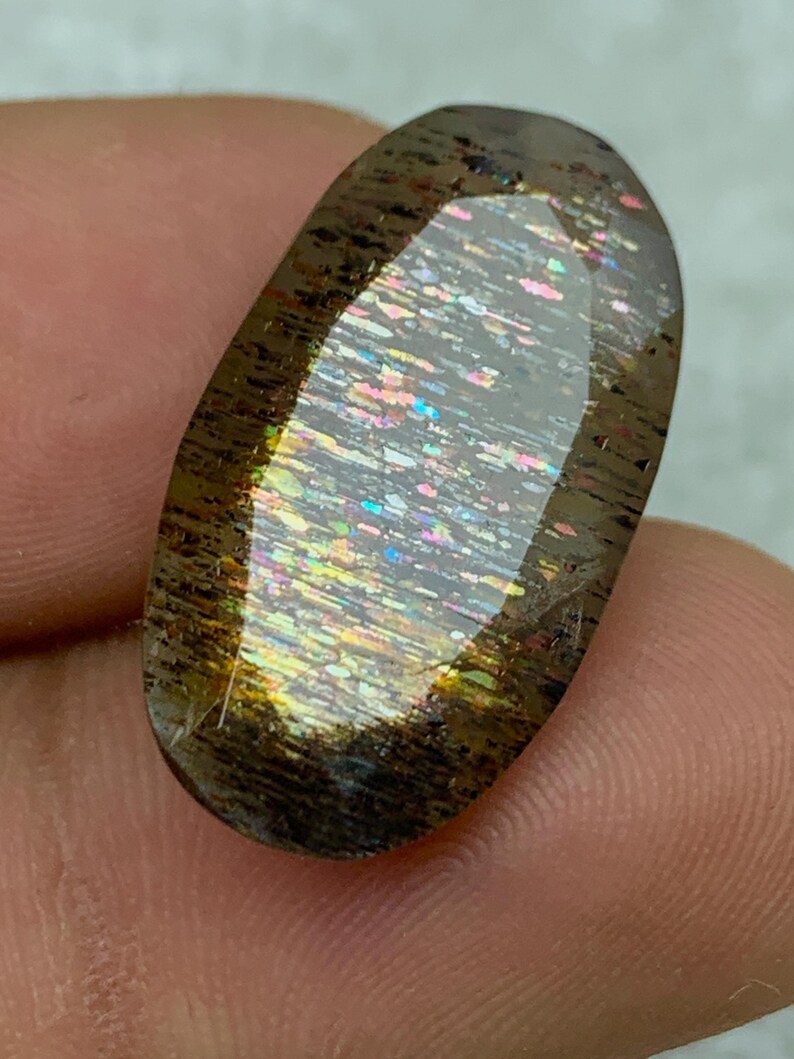 Op de afbeelding: Een grote, ovale edelsteen met een donkerbruine basis en een iriserende regenboogkleurige schittering. De edelsteen heeft een glad, gepolijst oppervlak.
