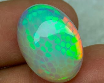 May include: An oval-shaped opal gemstone with a vibrant play of color. The stone displays a mosaic-like pattern with flashes of green, orange, and blue. The opal is held between two fingers, showcasing its polished surface and iridescent qualities.