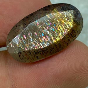Op de afbeelding: Een grote, ovale edelsteen met een doorschijnende, iriserende afwerking. De steen heeft een bruine basis met gouden, zilveren en regenboogkleurige flitsen.