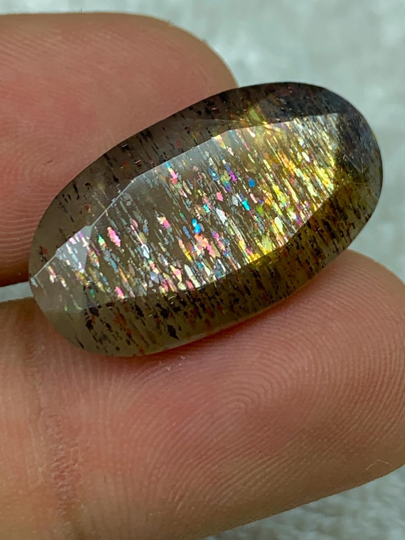 Op de afbeelding: Een grote, ovale edelsteen met een doorschijnende, iriserende afwerking. De steen heeft een bruine basis met gouden, zilveren en regenboogkleurige flitsen.
