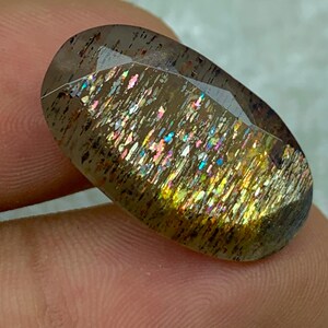 Op de afbeelding: Een grote, ovale, iriserende edelsteen met een bruine, gouden en regenboogkleurige glans. De edelsteen is gezet in een heldere, gefacetteerde zetting.