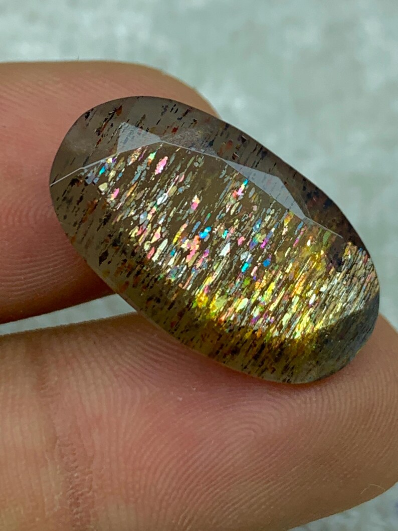 Op de afbeelding: Een grote, ovale, iriserende edelsteen met een bruine, gouden en regenboogkleurige glans. De edelsteen is gezet in een heldere, gefacetteerde zetting.