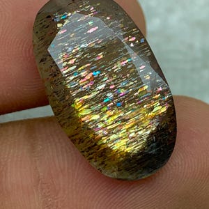 Op de afbeelding: Een grote, ovale edelsteen met een donkerbruine basis en een iriserende regenboogkleurige glans. De edelsteen heeft een glad, gepolijst oppervlak en is gezet in een heldere, transparante zetting.