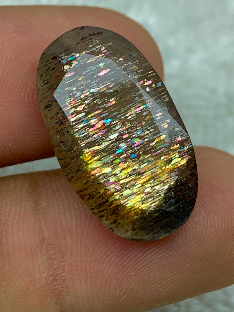 Op de afbeelding: Een grote, ovale edelsteen met een donkerbruine basis en een iriserende regenboogkleurige glans. De edelsteen heeft een glad, gepolijst oppervlak en is gezet in een heldere, transparante zetting.