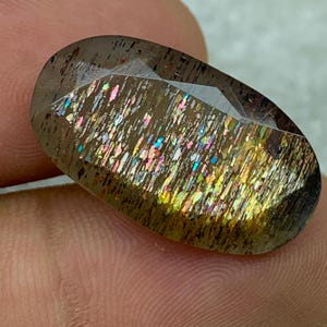 Op de afbeelding: Een grote, ovale edelsteen met een donkerbruine basis en iriserende regenboogkleuren. De edelsteen heeft een glad, gepolijst oppervlak en is gezet in een bezel-setting.