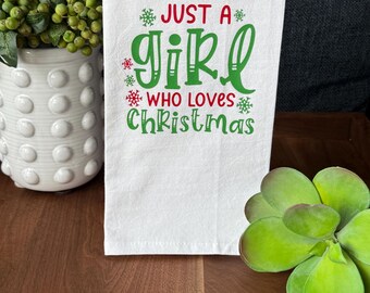May include: White kitchen towel with the text "JUST A GIRL WHO LOVES Christmas" in red and green. The towel is hanging on a black bar. A white vase with green berries and a green succulent are in the background.