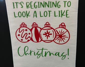 May include: White kitchen towel with the text "It's beginning to look a lot like Christmas!" in green lettering. Four red Christmas ornaments are depicted below the text.