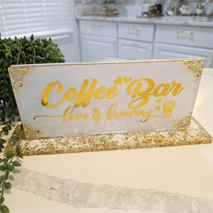 K&ouml;nnte beinhalten: Ein rechteckiges Schild aus wei&szlig;em Harz mit Blattgoldakzenten an den Ecken und am Sockel. Das Schild tr&auml;gt den goldenen Schriftzug Coffee Bar und love is brewing mit zwei kleinen Herzen und einem Kaffeetassensymbol. Es steht auf einem passenden Sockel.