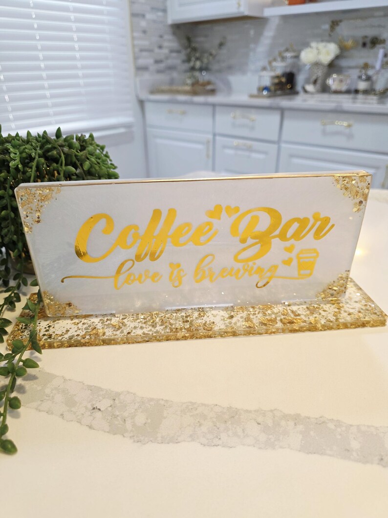 K&ouml;nnte beinhalten: Ein rechteckiges Schild aus wei&szlig;em Harz mit Blattgoldakzenten an den Ecken und am Sockel. Das Schild tr&auml;gt den goldenen Schriftzug Coffee Bar und love is brewing mit zwei kleinen Herzen und einem Kaffeetassensymbol. Es steht auf einem passenden Sockel.