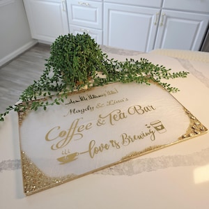 May include: A rectangular white sign with gold glitter edges and gold lettering that reads "Coffee & Tea Bar" and "love is brewing". A small green plant sits above the sign.