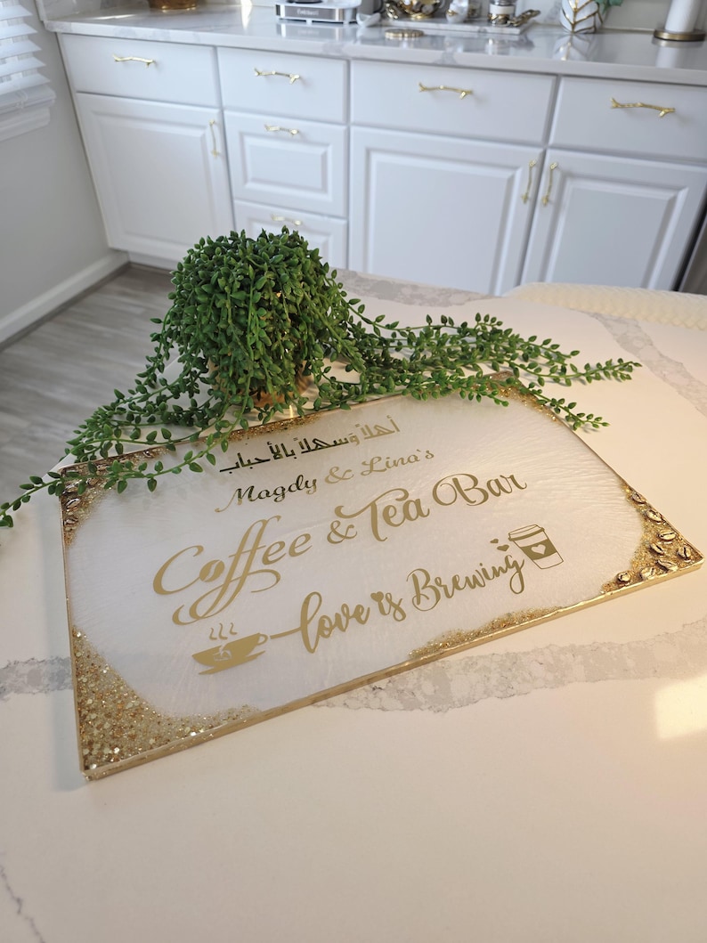 May include: A rectangular white sign with gold glitter edges and gold lettering that reads "Coffee & Tea Bar" and "love is brewing". A small green plant sits above the sign.