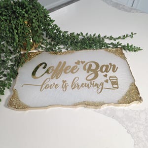 May include: A rectangular white coffee bar sign with gold glitter edging. The sign features the words "Coffee Bar love is brewing" in gold script, along with a coffee cup icon. Green leafy plant in the background.