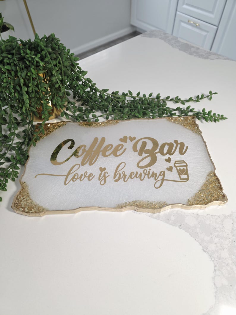 May include: A rectangular white coffee bar sign with gold glitter edging. The sign features the words "Coffee Bar love is brewing" in gold script, along with a coffee cup icon. Green leafy plant in the background.