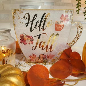 K&ouml;nnte beinhalten: Ein dekoratives, &uuml;bergro&szlig;es Teetassen-f&ouml;rmiges Schild mit den Worten "Hello Fall" in goldener Schrift. Das Schild hat einen wei&szlig;en Hintergrund mit Goldflocken, Herbstbl&auml;ttern und K&uuml;rbis-Illustrationen.