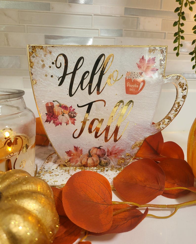 K&ouml;nnte beinhalten: Ein dekoratives, &uuml;bergro&szlig;es Teetassen-f&ouml;rmiges Schild mit den Worten "Hello Fall" in goldener Schrift. Das Schild hat einen wei&szlig;en Hintergrund mit Goldflocken, Herbstbl&auml;ttern und K&uuml;rbis-Illustrationen.