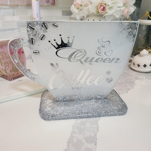 K&ouml;nnte beinhalten: Ein dekoratives Tablett in Form einer Kaffeetasse mit silberner Glitzerbasis und -rand. Die Tasse tr&auml;gt die Aufschrift "Queen Coffee" und ein Kronen-Design. Kaffeebohnen- und Herz-Akzente sind ebenfalls sichtbar.