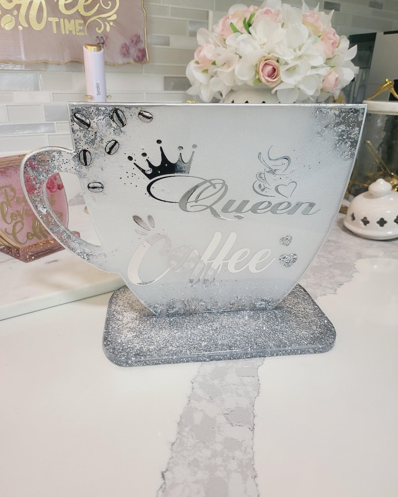K&ouml;nnte beinhalten: Ein dekoratives Tablett in Form einer Kaffeetasse mit silberner Glitzerbasis und -rand. Die Tasse tr&auml;gt die Aufschrift "Queen Coffee" und ein Kronen-Design. Kaffeebohnen- und Herz-Akzente sind ebenfalls sichtbar.