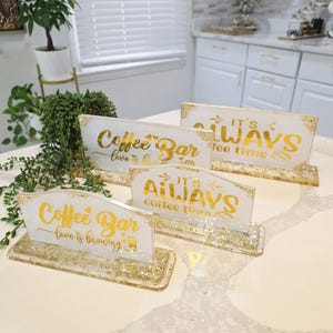 K&ouml;nnte beinhalten: Vier rechteckige Harzschilder mit goldglitzernden Sockeln und wei&szlig;en Oberfl&auml;chen. Zwei Schilder tragen den goldenen Schriftzug Coffee Bar love is brewing, zwei weitere den goldenen Schriftzug IT'S ALWAYS coffee time, auf einer wei&szlig;en Arbeitsplatte.