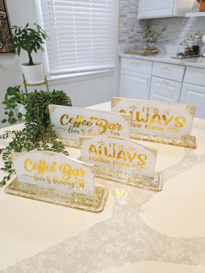 K&ouml;nnte beinhalten: Vier rechteckige Harzschilder mit goldglitzernden Sockeln und wei&szlig;en Oberfl&auml;chen. Zwei Schilder tragen den goldenen Schriftzug Coffee Bar love is brewing, zwei weitere den goldenen Schriftzug IT'S ALWAYS coffee time, auf einer wei&szlig;en Arbeitsplatte.