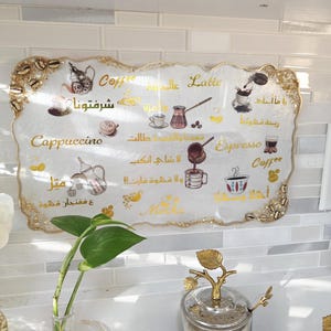 May include: A decorative coffee-themed sign with gold trim and Arabic text. The sign features illustrations of coffee drinks like cappuccino, espresso, and latte. The sign is white with gold accents and is displayed in a kitchen setting.