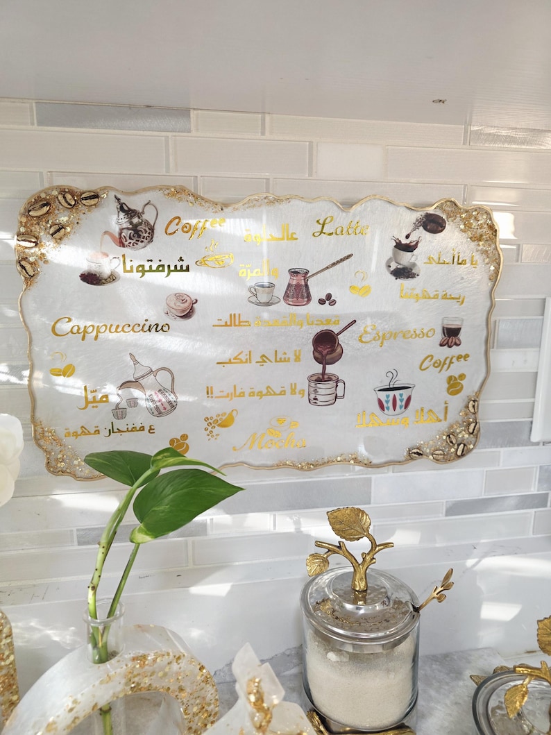 May include: A decorative coffee-themed sign with gold trim and Arabic text. The sign features illustrations of coffee drinks like cappuccino, espresso, and latte. The sign is white with gold accents and is displayed in a kitchen setting.