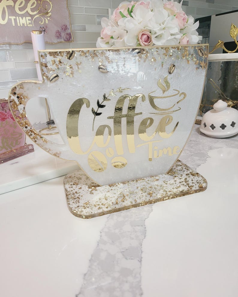 K&ouml;nnte beinhalten: Ein gro&szlig;er, dekorativer Halter in Form einer Kaffeetasse mit den Worten "Coffee Time" in goldfarbenen Buchstaben. Die Tasse ist wei&szlig; mit goldenen Akzenten und Kaffeebohnen-Verzierungen. Ein kleiner Strau&szlig; rosa und wei&szlig;er Blumen befindet sich im Inneren.
