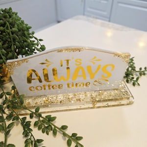 K&ouml;nnte beinhalten: Ein rechteckiges Schild aus wei&szlig;em Kunstharz mit eingearbeiteten Blattgoldflocken an der Basis und den Kanten. Das Schild tr&auml;gt den goldenen Schriftzug IT'S ALWAYS coffee time und eine kleine Kaffeebohne. Das St&uuml;ck steht auf einem passenden Sockel.