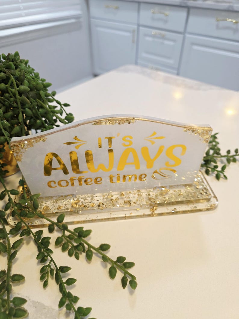 K&ouml;nnte beinhalten: Ein rechteckiges Schild aus wei&szlig;em Kunstharz mit eingearbeiteten Blattgoldflocken an der Basis und den Kanten. Das Schild tr&auml;gt den goldenen Schriftzug IT'S ALWAYS coffee time und eine kleine Kaffeebohne. Das St&uuml;ck steht auf einem passenden Sockel.
