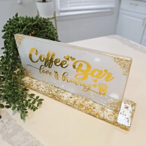 K&ouml;nnte beinhalten: Ein rechteckiges Schild aus wei&szlig;em Harz mit Blattgoldakzenten und einem goldenen St&auml;nder. Das Schild tr&auml;gt den Text Coffee Bar und love is brewing in goldener Schreibschrift, begleitet von kleinen Herzsymbolen und einer Kaffeetassengrafik.