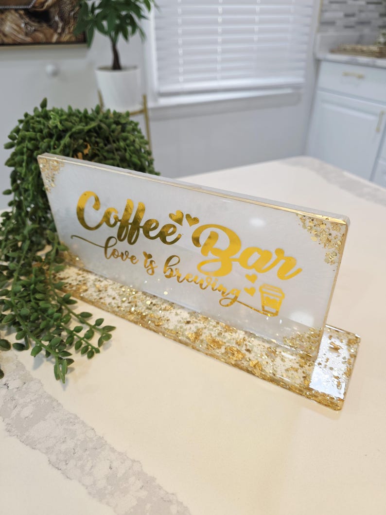 K&ouml;nnte beinhalten: Ein rechteckiges Schild aus wei&szlig;em Harz mit Blattgoldakzenten und einem goldenen St&auml;nder. Das Schild tr&auml;gt den Text Coffee Bar und love is brewing in goldener Schreibschrift, begleitet von kleinen Herzsymbolen und einer Kaffeetassengrafik.