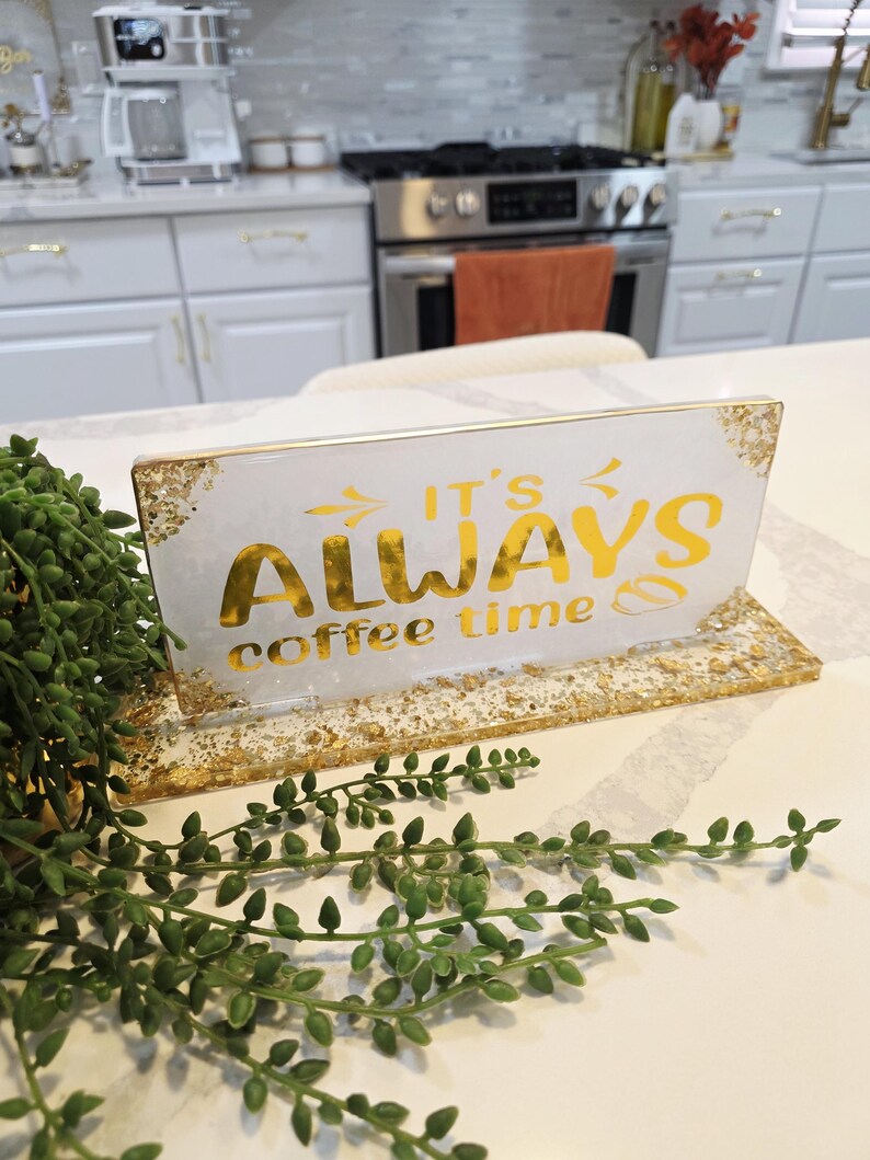 K&ouml;nnte beinhalten: Ein rechteckiges Schild aus wei&szlig;em Harz mit eingebetteten Blattgoldflocken an den Kanten und am Sockel. Das Schild tr&auml;gt den goldenen Schriftzug "IT'S ALWAYS coffee time" sowie kleine Pfeil- und Kaffeebohnengrafiken.