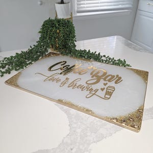 May include: A rectangular coffee bar serving tray with a white and gold design. The tray has gold glitter accents on the edges and corners, with the words "Coffee Bar love is brewing" in gold script. A coffee cup icon is also included.